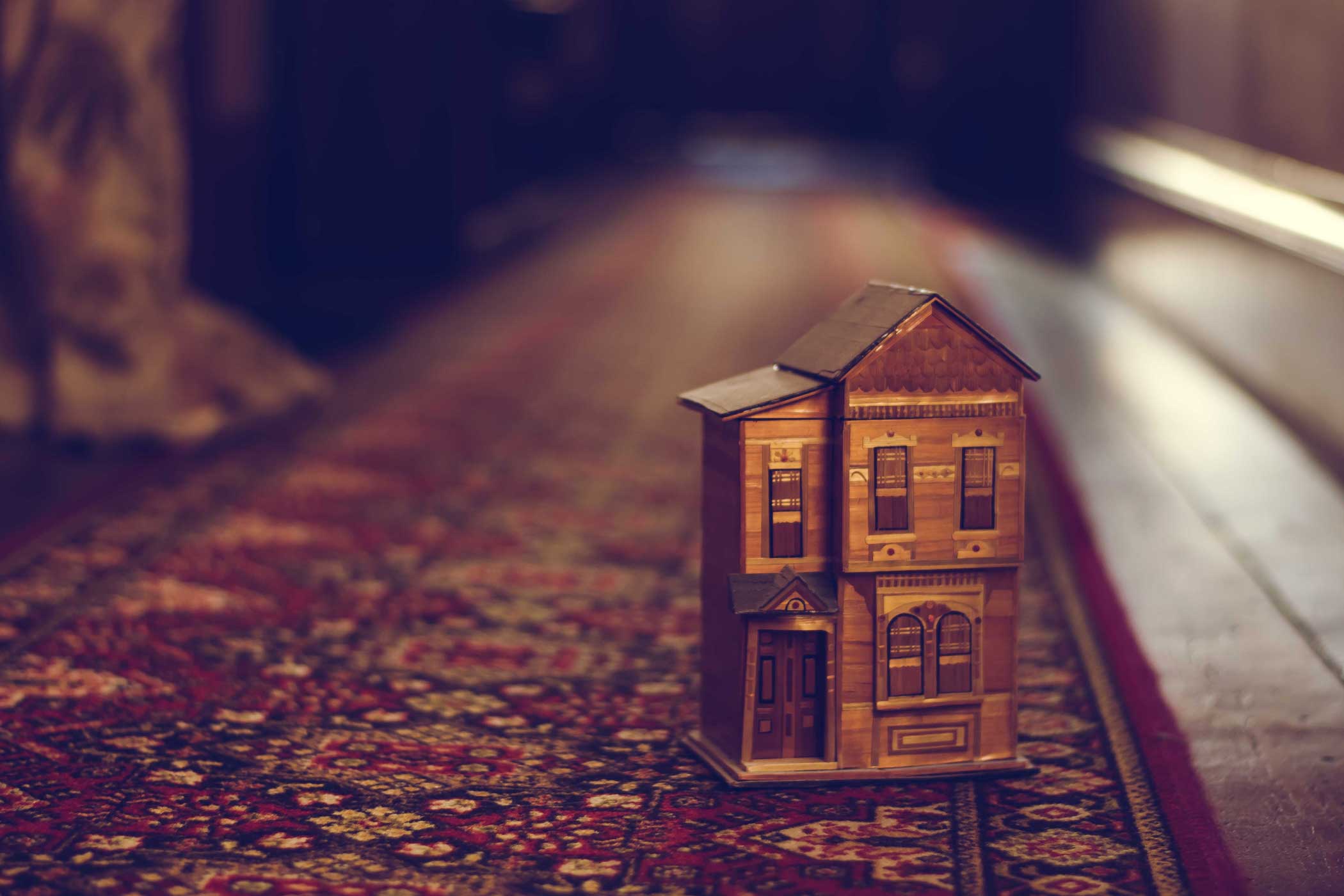 A close-up of a small model of a wood-framed town house sitting on a red patterned carpet on top of wooden floor boards.