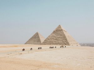 stone pyramids in desert, people and camels traveling on dirt road below