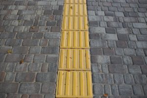 the picture shows a grey pavement. In the middle there are yellow warning tiles for visually impaired users.